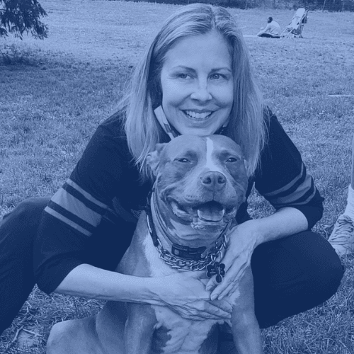 Photo of a smiling light-skinned woman with blond hair and a dark sweater standing behind a smiling pitbull terrier in a park