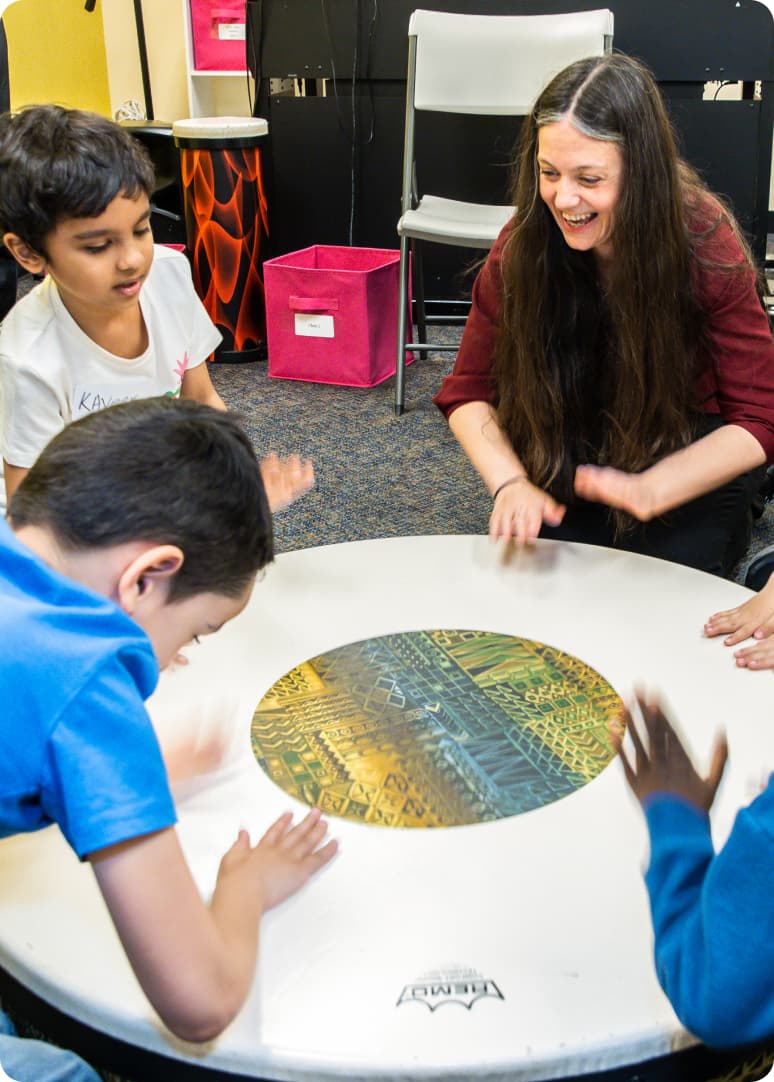 DMF Director of Teaching and Learning Juliana leads a circle of DMF participants playing a beat with their hands on a large drum