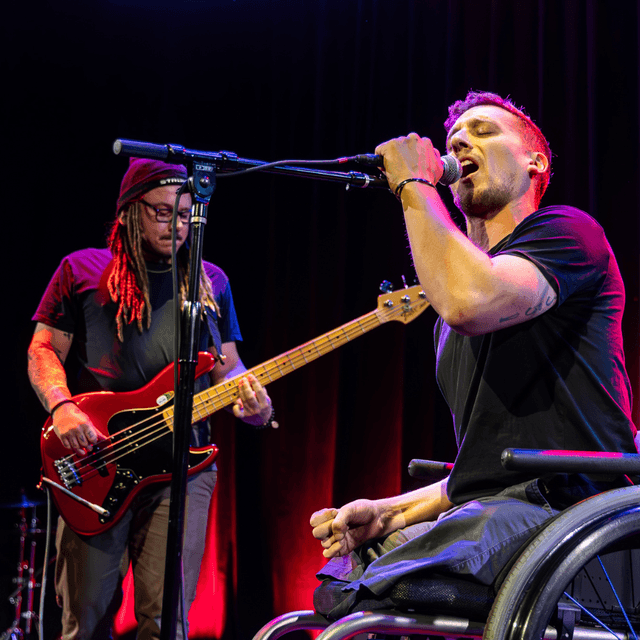 Photo of two members of the resilient. The front man is using a wheelchair, and singing into a microphone on the stand. Behind him is the bassist, with dreadlocks and a black beanie. They are both light skinned and in their early 30s.