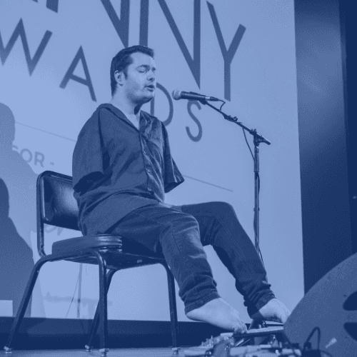 Photo of George playing guitar on stage. He is a caucasian man with brown hair and og average build without arms, playing guitar with his feet.