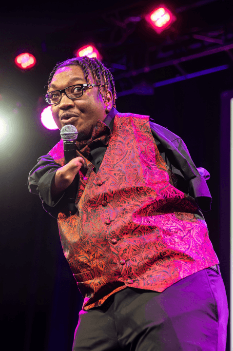 Photo of Gifted Hands singing at the 2024 Danny Awards. He has shortened arms, braided hair, glasses, and is wearing a red vest. 