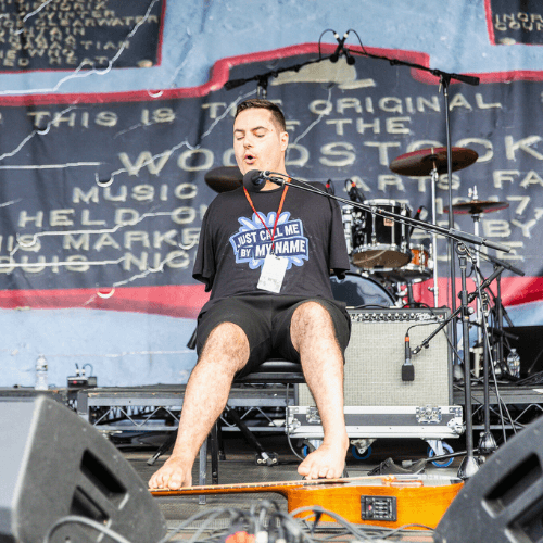 George Dennehy performing on stage at the Horizon Stage at Bethel Woods Center for The Arts. He is an armless man with short hair, of average height and build, and is playing guitar with his feet. 