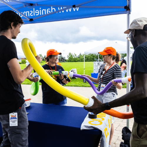 Photo of the accessible instrument table at Hello Inclusion, where two DMF staff members are assisting visitors in playing an instrument composed of four pool noodles