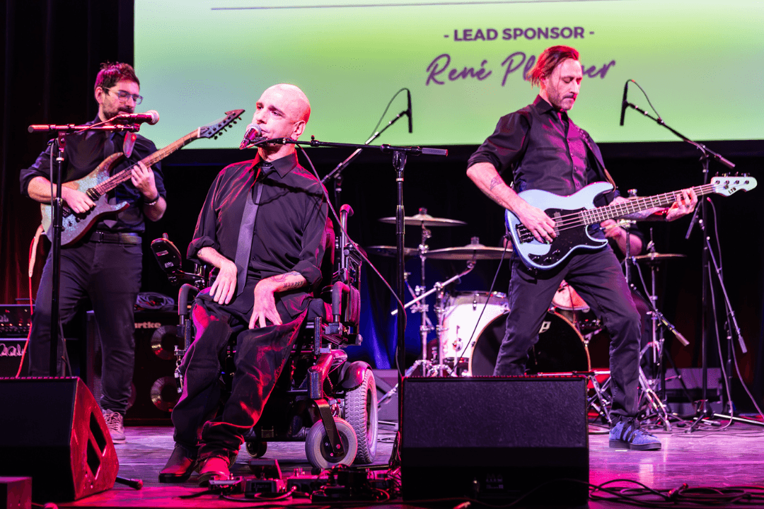 Photo of the band Corevalay performing at The Danny Awards, a four-piece featuring a drummer, guitarist, bassist, and singer, who is using a wheelchair, centered at the front of the stage. 