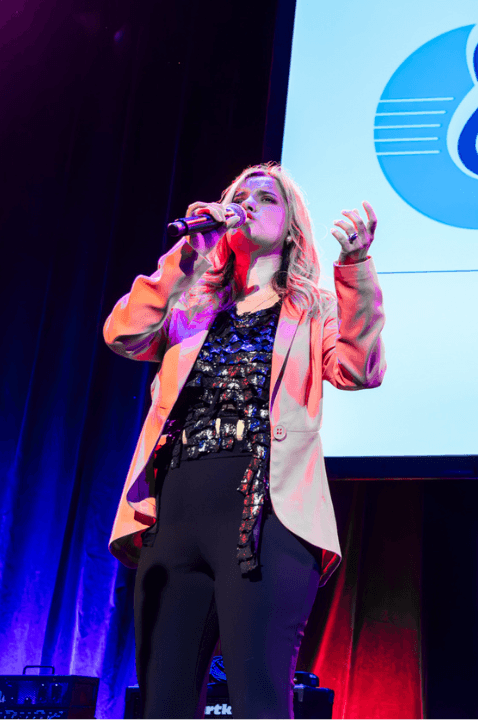 Photo of Rachael Leahcar singing at the 2024 Danny Awards. She has blond hair, and is dressed in black, with a peach jacket.  