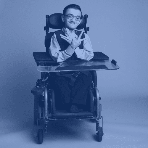 Photo of a young south Asian man using a wheelchair. He has slicked back hair and is wearing glasses. He is smiling and making hand signs.