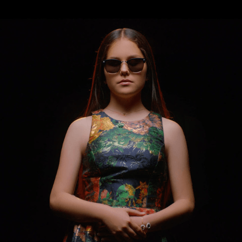 Photo of Iolanta. A young white woman with long, straight brown hair. She is wearing a colorful dress and dark glasses, and is standing in front of a black background.