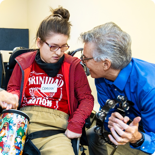 DMF Artistic Director Gerry plays a tambourine and smiles at a DMF participant banging a drum