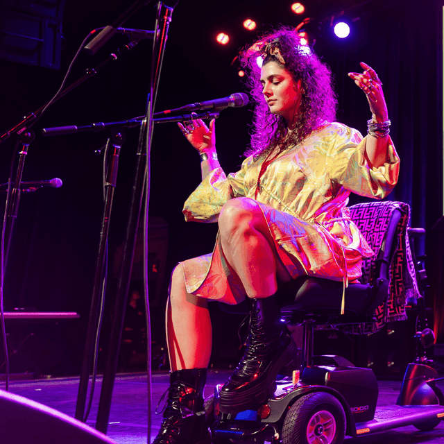 Photo of Ruth Lyon performing on stage at the Danny Awards. She is a white woman with long curly hair, wearing a light colored dress, while sitting in a wheelchair and singing into a microphone on a stand