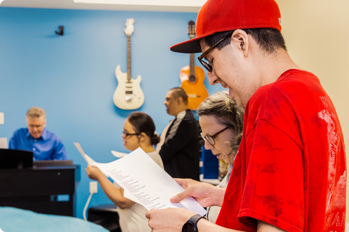 4 DMF participants sing a song in the colorful music center. In the background, DMF Artistic Director Gerry plays the piano. Guitars hang on the wall